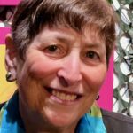Closeup photo of a woman smiling at the camera with a colorful background and plants behind her.