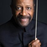 Closeup headshot of a man smiling and holding a music conducting baton.