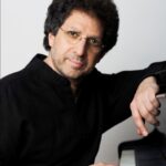 Headshot of a man sitting at a piano with one hand resting on the keys as he looks at the camera.