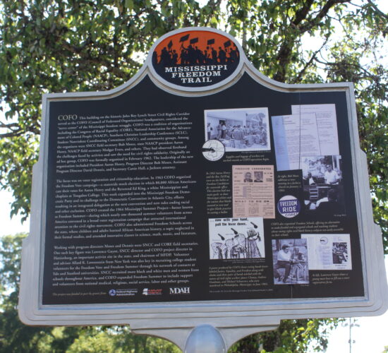 A historical marker displays paragraphs of text describing the history of the COFO building. To the right of the paragraphs is a collage of photos and items pertaining to COFO. Trees stand behind the marker.