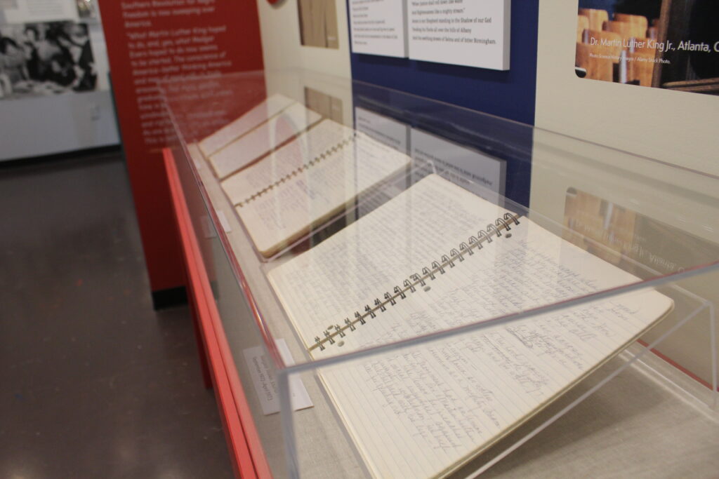 A series of documents are enclosed in a clear display case in front of an exhibit wall.