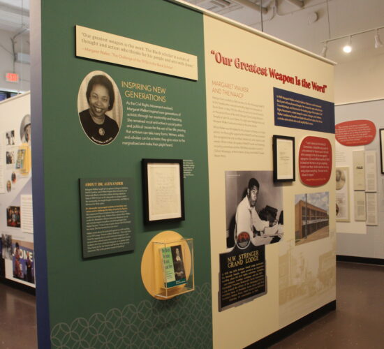 The walls of an exhibit partition a room in half and are filled with photos, blocks of text, framed documents, and more.