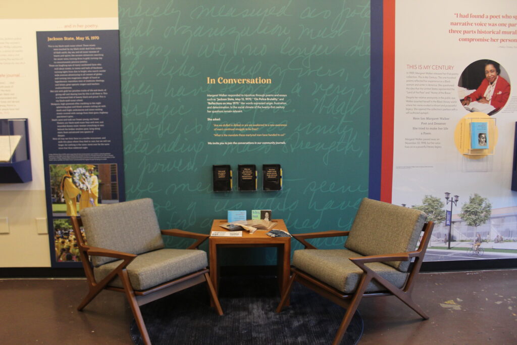 Two chairs are positioned around a table in front of an exhibit wall with text on it. The table is filled with books, bookmarks, and other materials. On the wall in holders are three notebooks.