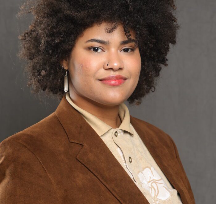 Professional headshot of a person wearing a brown blazer, smiling against a dark beige background.