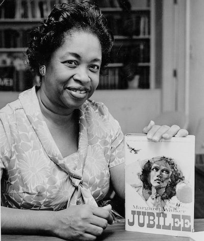 11Margaret Walker smiles while holding a copy of her novel Jubilee upright on a table. The cover shows an illustration of a woman looking upward.