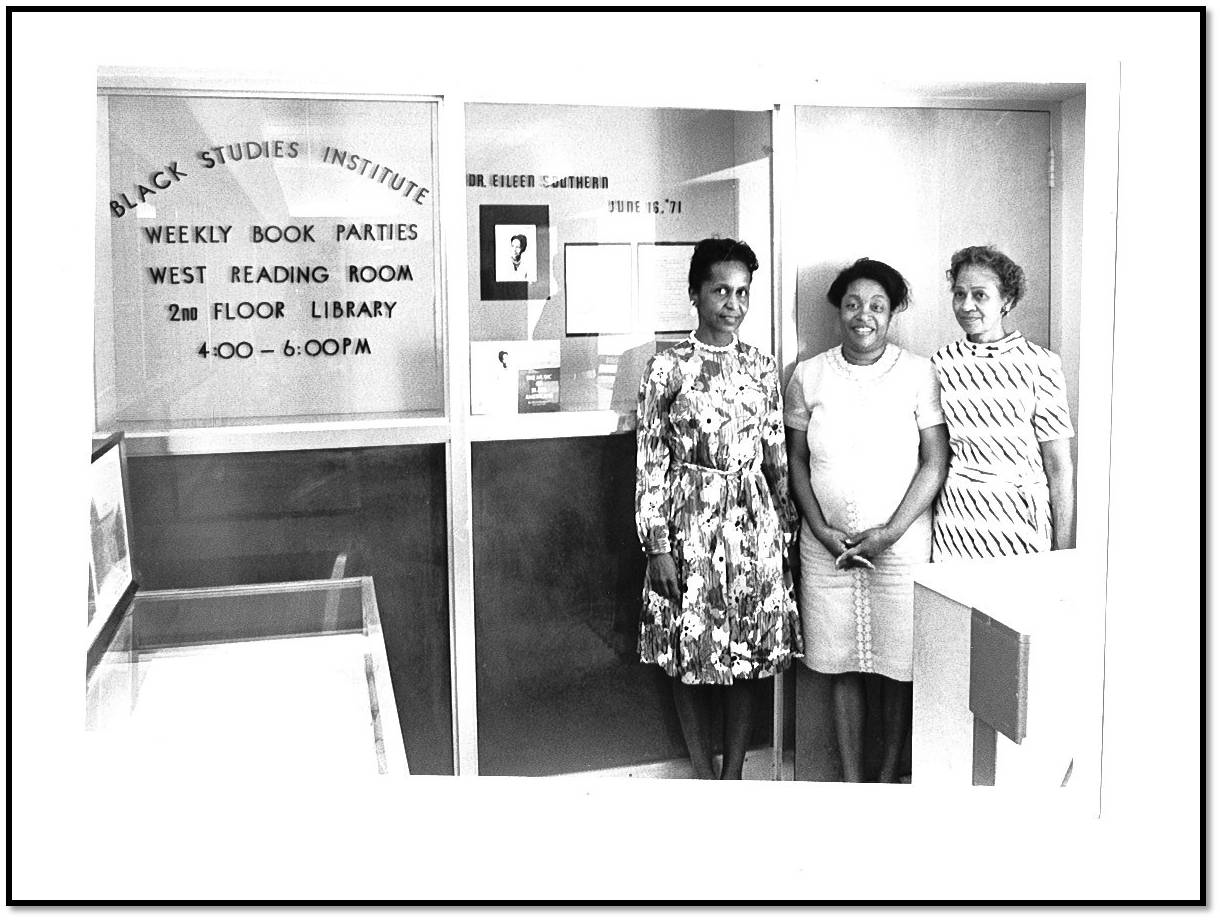 Three women stand outside a room with glass windows. On the wall inside the room is a sign advertising weekly reading parties with the Black Studies Institute.