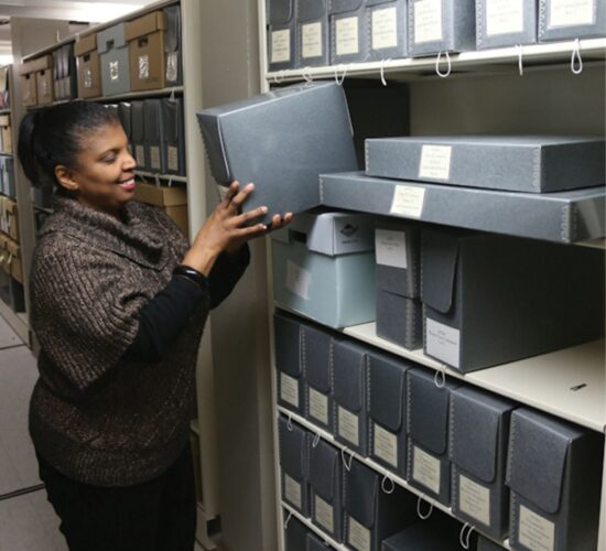 A woman smiles while putting a blue archival box on a shelf with many other boxes of varying sizes. There are other bookshelves in the background.